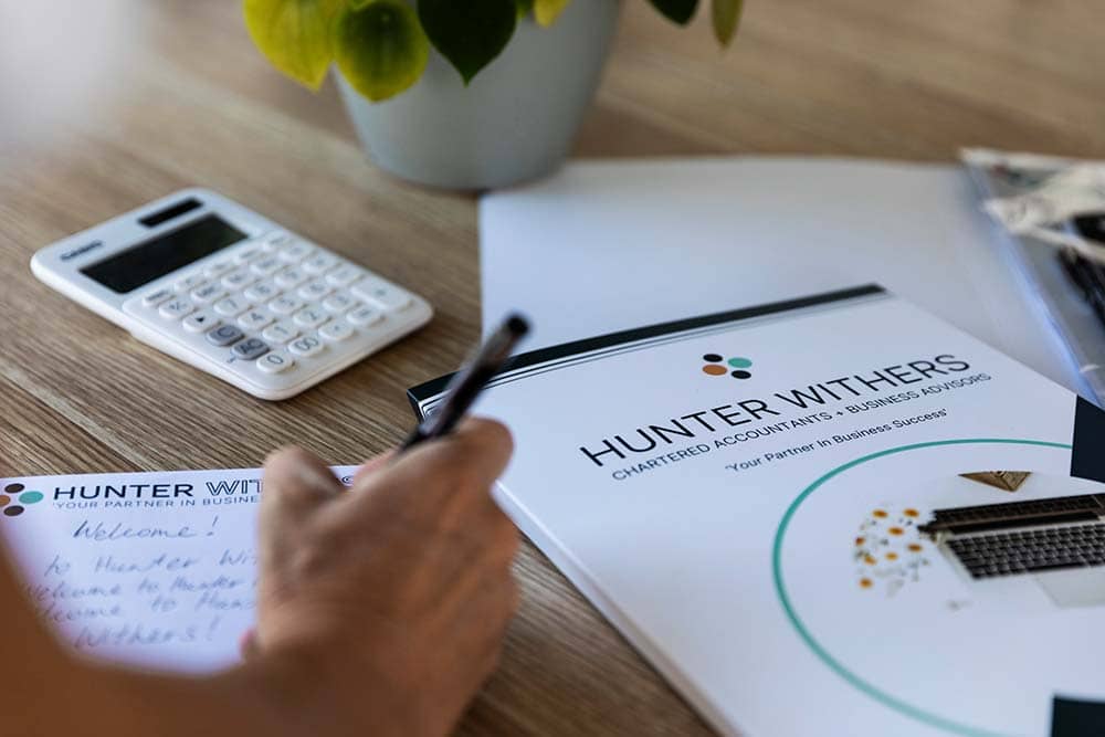 Close-up of a Hunter Withers branded folder, calculator, and a team member writing notes during a meeting at the Pukekohe office.