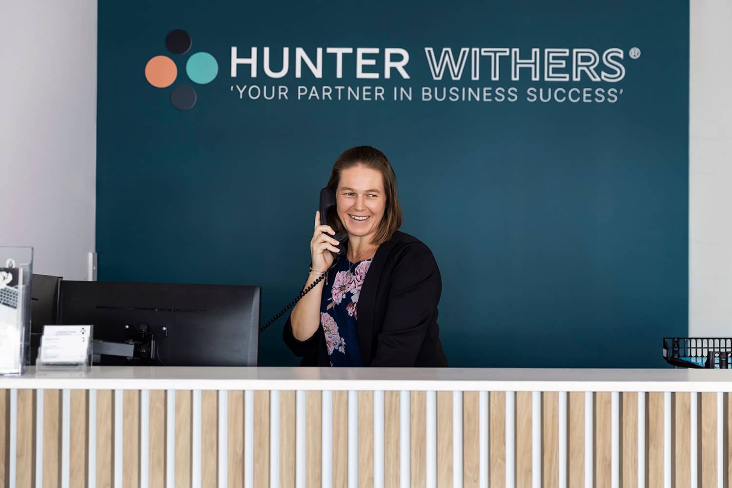 Cherie Webster, Admin Leader at Hunter Withers, seated at the front reception desk with branded wall in the Pukekohe office.