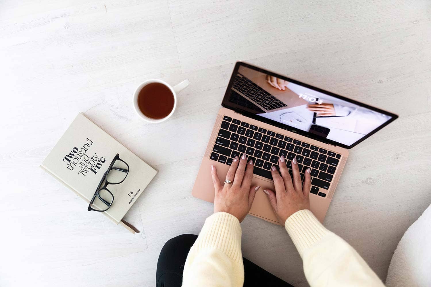 Top-down view of Emma Bracken working on her laptop, with a hot drink, 2025 planner, and reading glasses arranged nearby, capturing a focused and inviting workspace.