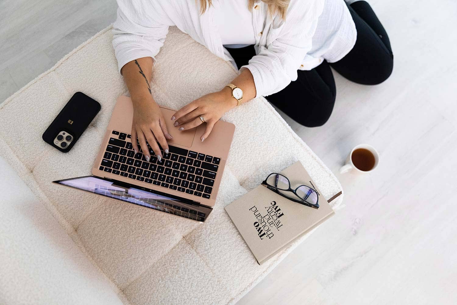 op-down view of Emma Bracken working from a couch on her laptop, with a phone, 2025 planner, hot drink, and reading glasses thoughtfully arranged nearby.