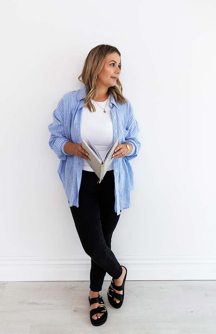 Full-length image of financial advisor Emma Bracken gazing thoughtfully out the window while holding her 2025 planning diary, dressed professionally in a light-filled office space.