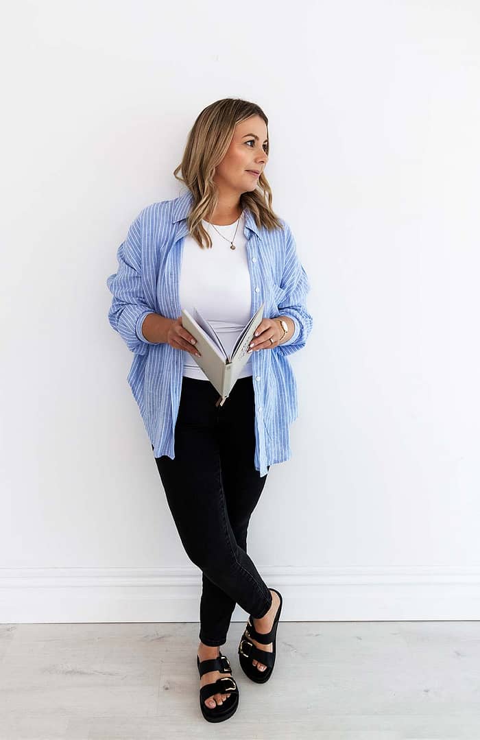 Full-length image of financial advisor Emma Bracken gazing thoughtfully out the window while holding her 2025 planning diary, dressed professionally in a light-filled office space.