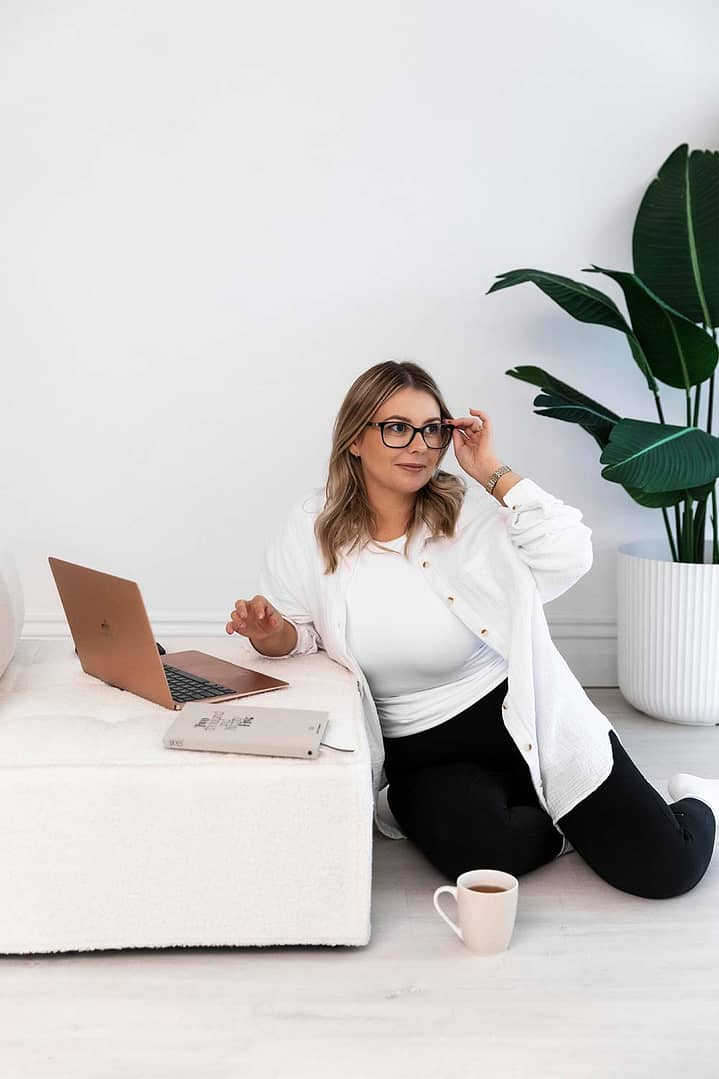 Emma Bracken working from the living room floor with her laptop open, playfully glancing out the window while adjusting her glasses - blending ease, focus, and personality.