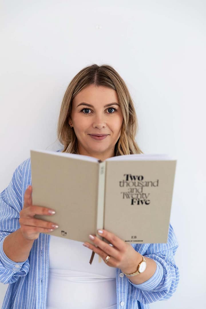 Emma Bracken, a financial advisor, smiling warmly at the camera while holding up a 2025 planning diary in a bright, professional setting.
