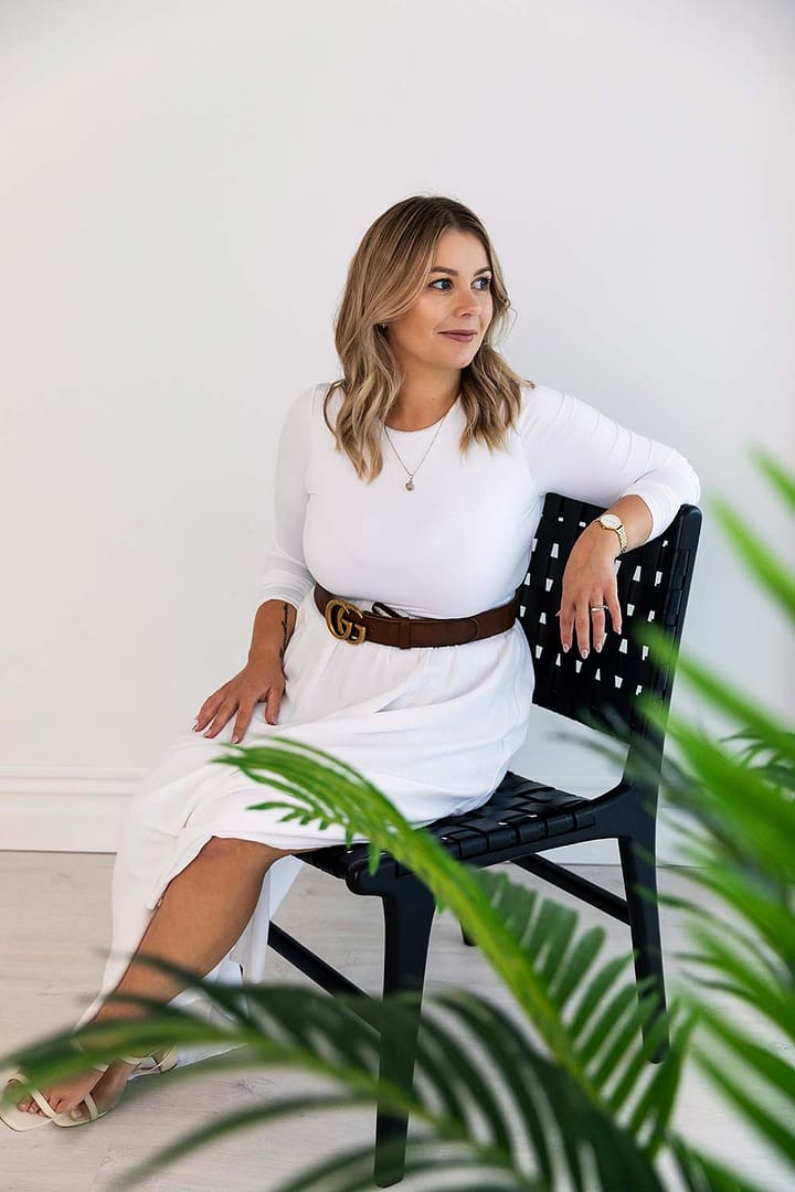 Emma Bracken, financial advisor, seated on a chair and leaning slightly to look over the backrest toward a window, softly framed by out-of-focus greenery in the foreground.